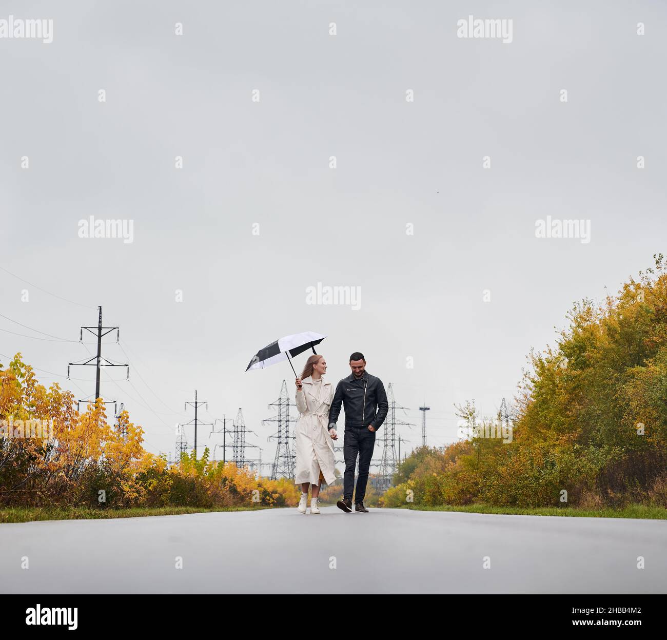 Un couple souriant et heureux marchant sur une route asphaltée tenant les bras.Fille regardant son amant, tenant un parapluie dans une main.En arrière-plan, les tours électriques et le ciel gris, les buissons jaunes des deux côtés de la route. Banque D'Images