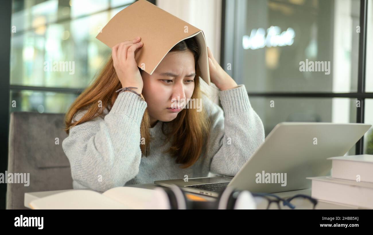 Une adolescente asiatique tenant un livre sur sa tête a stressé regardant l'ordinateur portable pendant les leçons en ligne de l'ordinateur portable, elle étudie en ligne à la maison. Banque D'Images