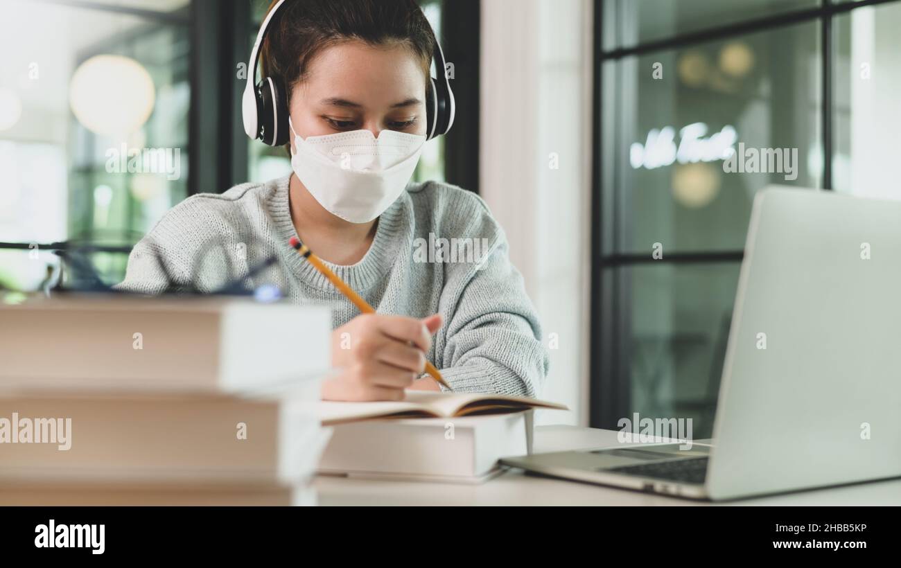 Fille asiatique avec stéthoscope portant un masque médical prenant des notes et étudiant en ligne avec un ordinateur portable. Banque D'Images