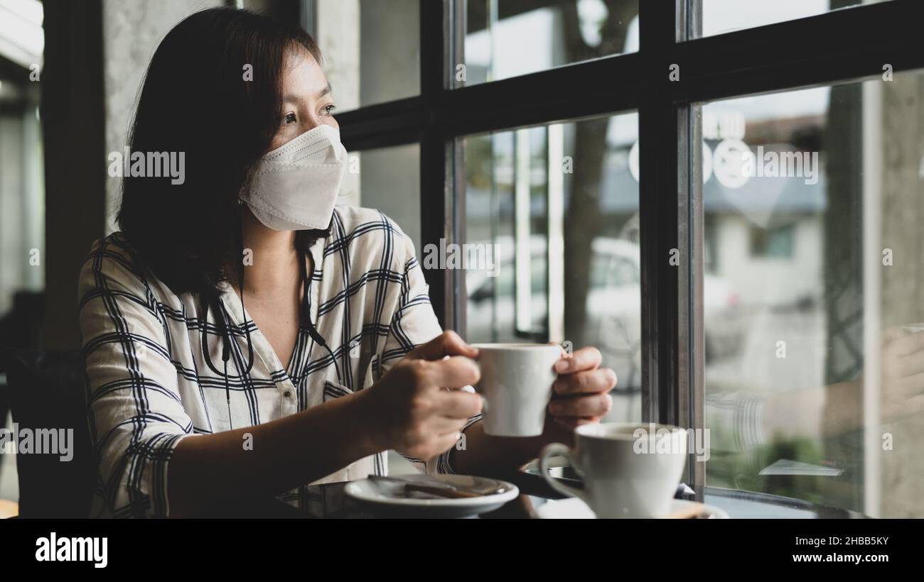 Une femme portant un masque médical est assise dans un café en regardant par la fenêtre avec des yeux tristes, elle est nourrie de l'épidémie de virus et veut vivre une n Banque D'Images
