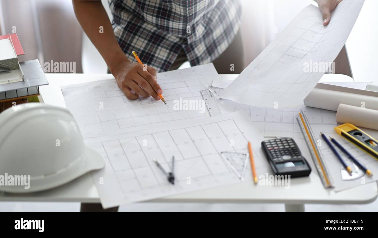 Homme de designer tenant un crayon examinant les plans de maison, casque de sécurité et maisons modèles avec des outils de dessin sur un bureau, architecte de conception de maison. Banque D'Images