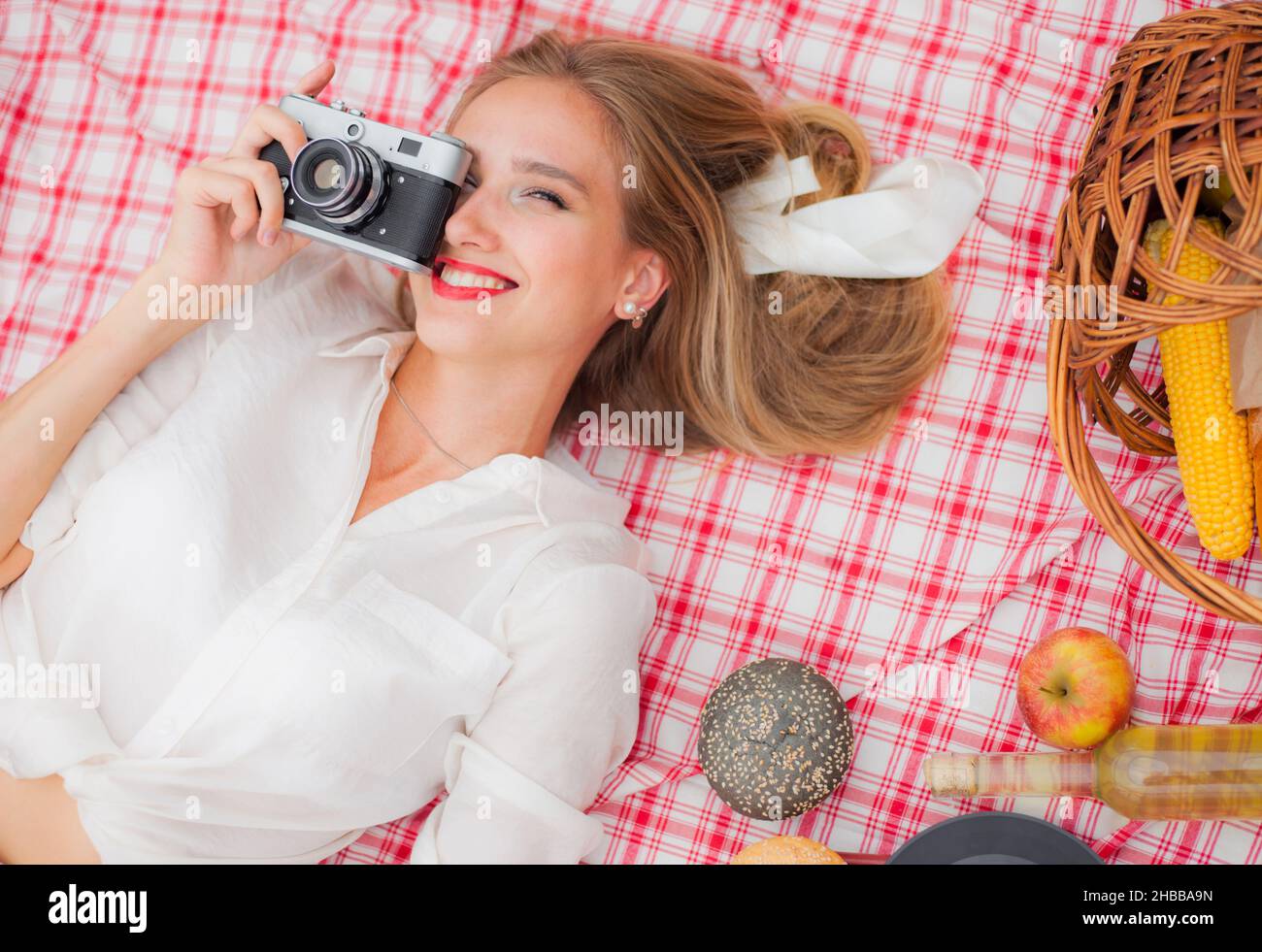 Pique-nique de style rétro.Jeune femme blonde gaie vêtue de vêtements vintage tenant un appareil photo rétro et prenant des photos tout en se reposant sur une table de pique-nique à l'extérieur.Haut Banque D'Images