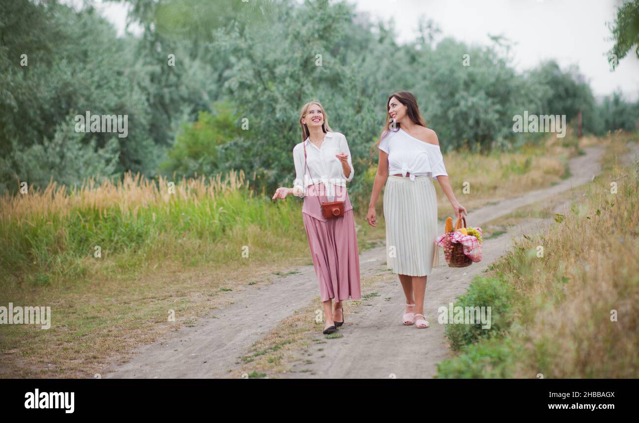 Deux jeunes femmes gaies en vêtements rétro marchent le long de l'atterrissage avec un panier de pique-nique et parlent.Promenade conjointe dans la nature Banque D'Images