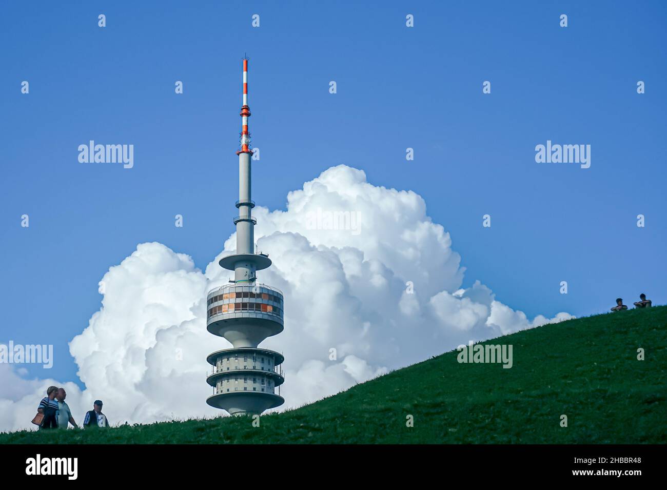 Un couple et un homme marchent sur une colline près de la Tour Olympique de Munich.Deux jeunes hommes s'assoient en haut à droite.Grands nuages dans le ciel bleu. Banque D'Images