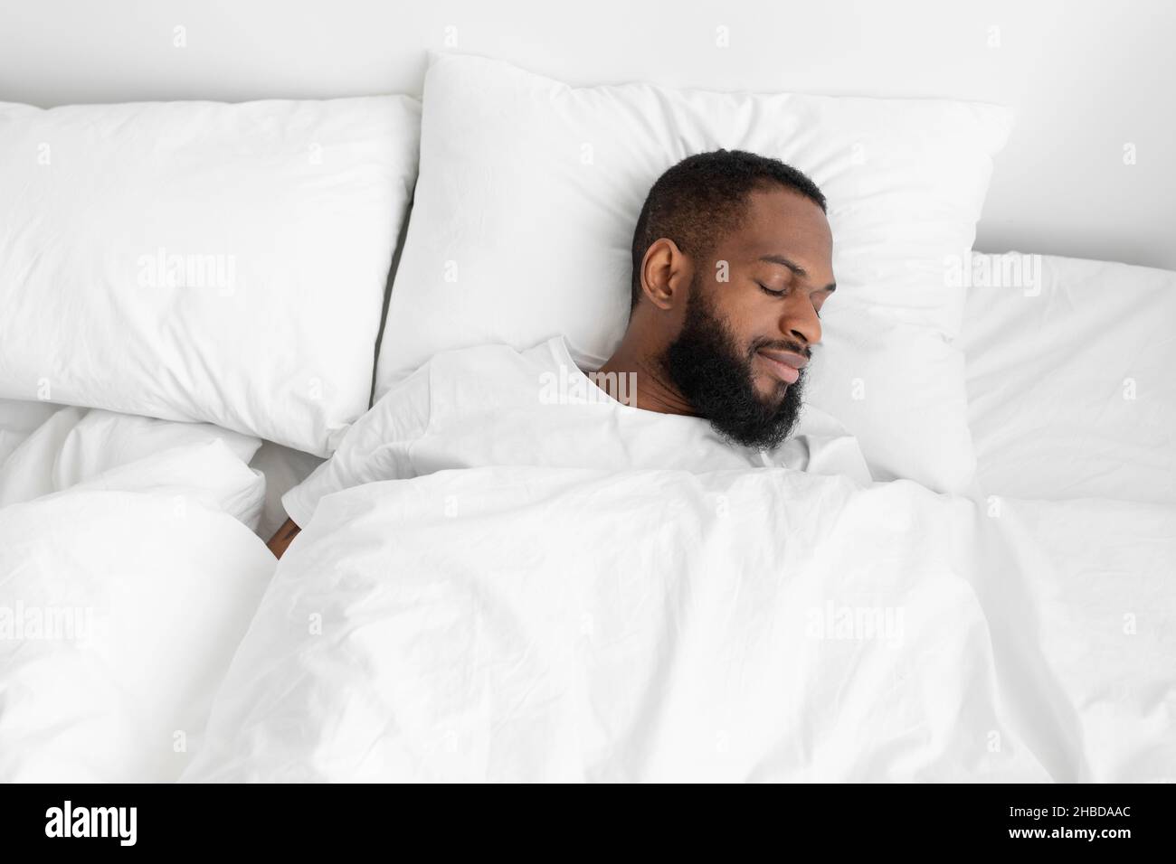 Calme millénaire noir barbu homme dormir sur le lit, oreiller doux avec couette blanche dans la chambre profiter du repos Banque D'Images