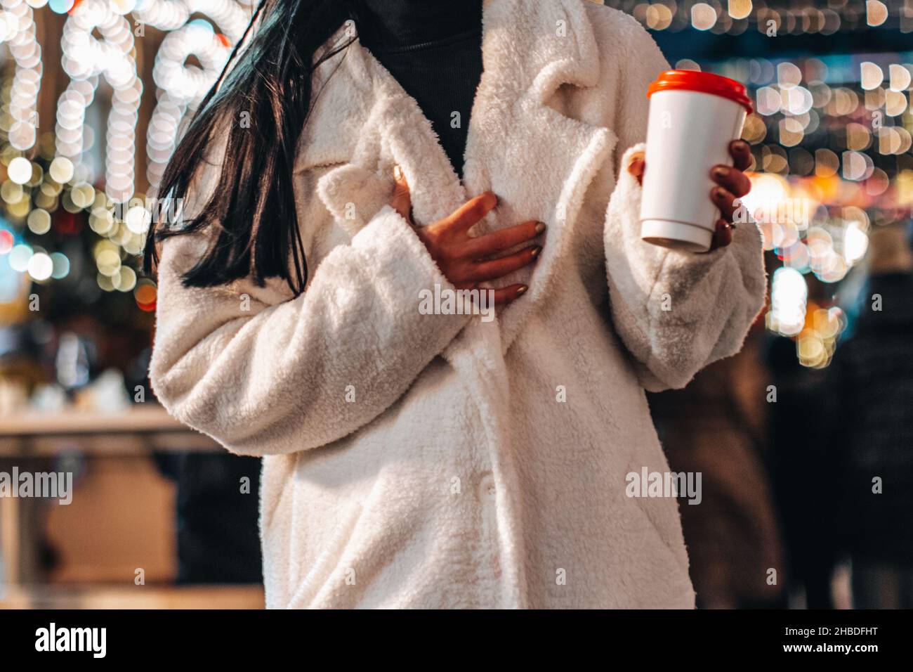 Corps féminin court en hiver chaud manteau de fourrure blanc tenant une tasse de plastique avec thé chaud ou café sur le fond de lumières de guirlande de bokeh.Christm Banque D'Images