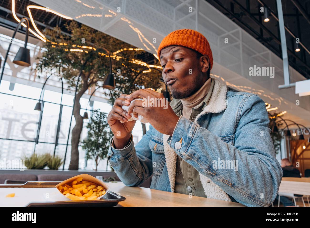 Un homme africain dans un chapeau orange et une veste en denim mange un délicieux hamburger et des frites dans un centre commercial sur une aire de restauration, des fast-food gras et malsains Banque D'Images