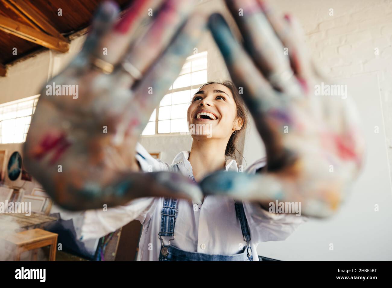 Une jeune peintre heureuse cadrage de son visage avec ses mains.Femme peintre insouciante regardant avec un sourire tout en se tenant dans son studio d'art.Joyeux jeune va Banque D'Images