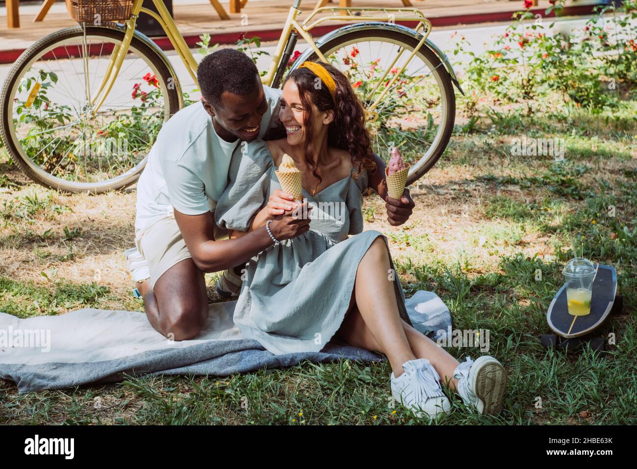 Un jeune homme afro-américain et une brunette s'assoient sur un couvre-lit sur l'herbe en mangeant de la glace et en riant le jour d'été ensoleillé dans le parc Banque D'Images