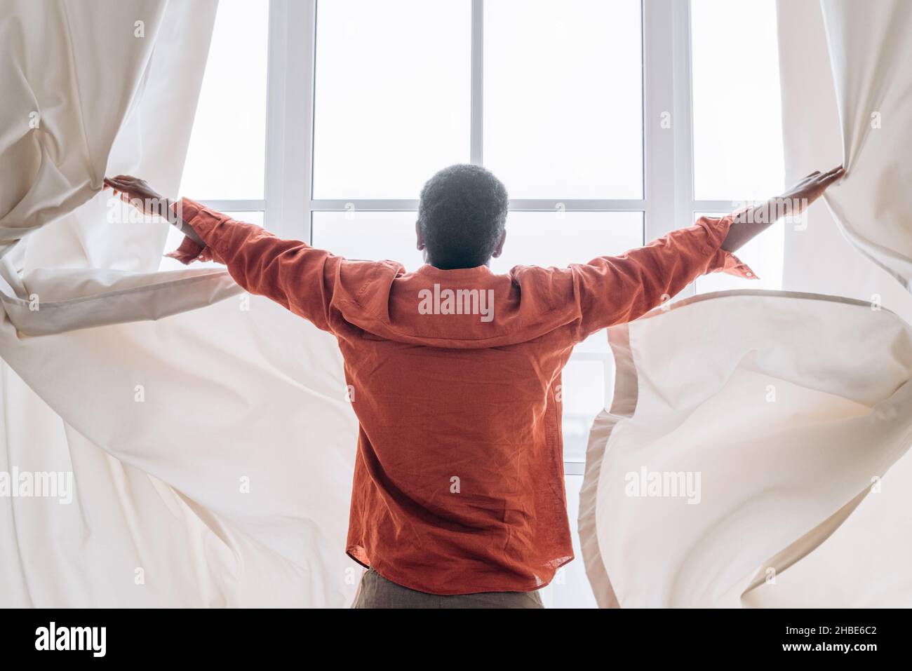 Un homme africain en pyjama orange ouvre les rideaux dans sa chambre après s'être réveillé le matin debout à une fenêtre lumineuse Banque D'Images