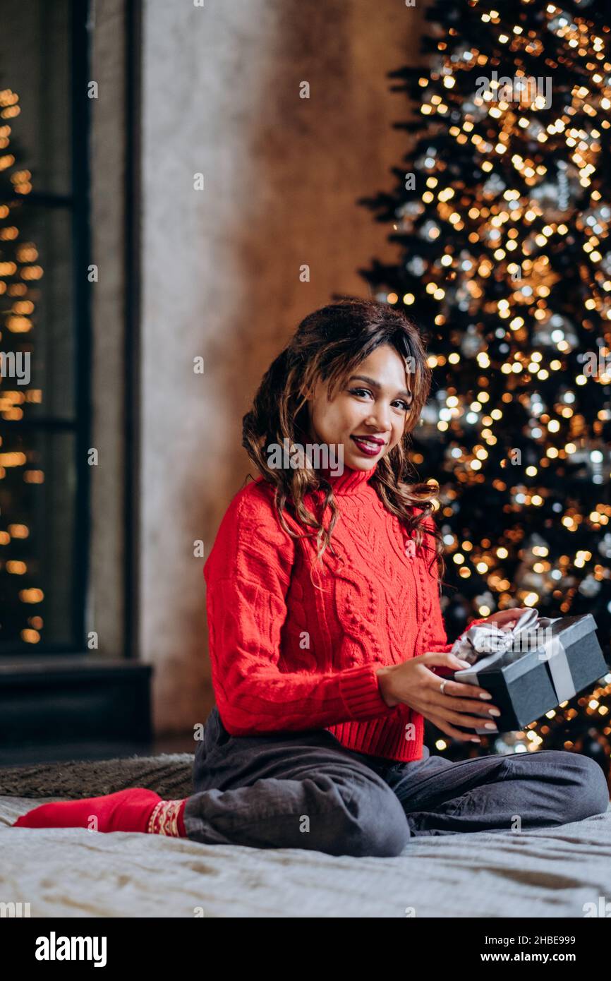 belle femme de course mixte dans un chandail rouge s'assoit sur le lit et tient un cadeau sur le fond d'un arbre de noël dans un studio loft, une femme sourit à la photo verticale de l'appareil photo Banque D'Images