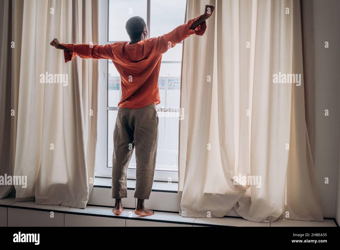 L'homme africain en pyjama et pantalon orange s'étire après s'être réveillé debout à la fenêtre avec des rideaux et regarde dans la distance, faisant des exercices matinaux, le début d'une nouvelle journée Banque D'Images