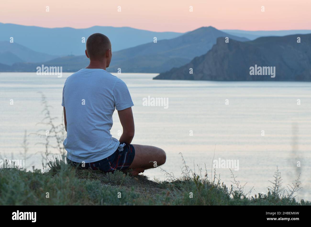 L'homme s'assoit sur une falaise au-dessus de la mer et regarde le coucher du soleil.Soirée chaude sur la côte Banque D'Images