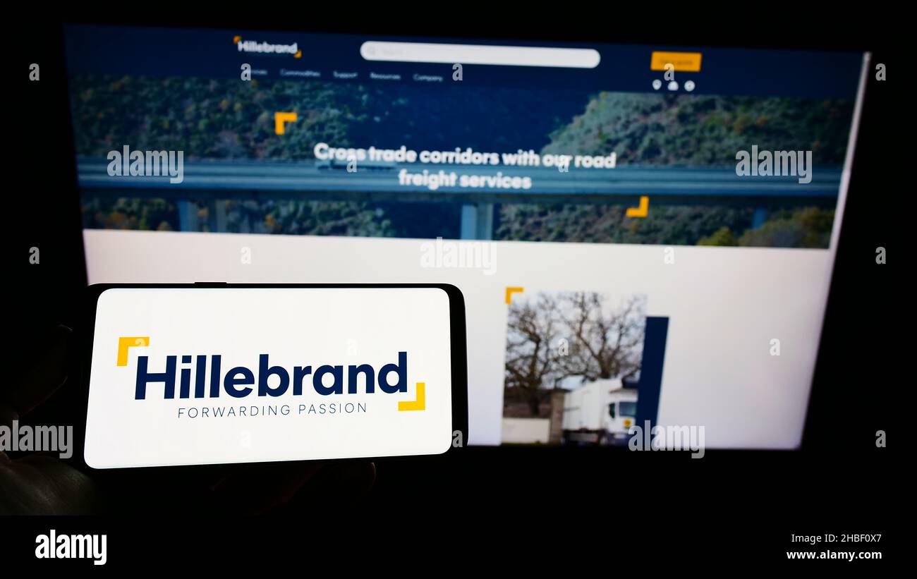 Personne portant un smartphone portant le logo de la société allemande de logistique J.F.Hillebrand Group AG à l'écran en face du site Web.Mise au point sur l'affichage du téléphone. Banque D'Images