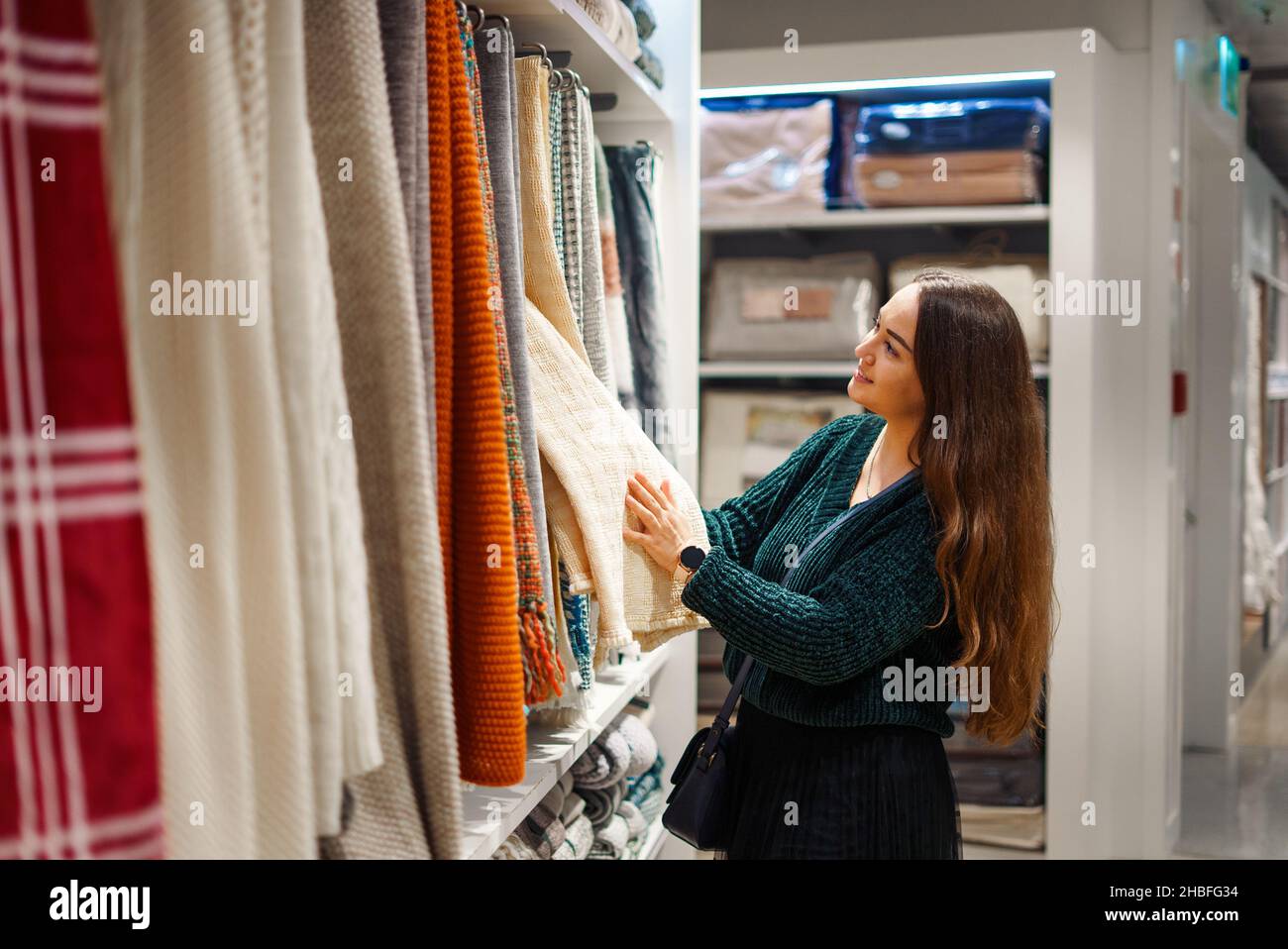 Une femme souriante qui fait du shopping pour sa nouvelle maison, qui se trouve dans le magasin de produits ménagers, regarde sur un stand avec des couvertures suspendues de différentes couleurs, touche du tissu, évalue la qualité des produits Banque D'Images