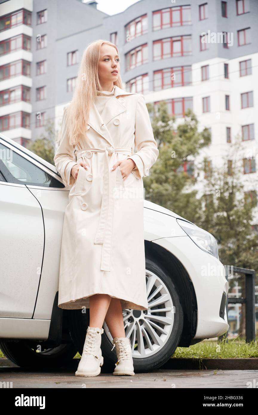 Vue basse d'une fille blonde dans un manteau blanc appuyé contre sa voiture moderne à l'extérieur sur fond d'arbre et de bâtiment en hauteur.Dame debout près de l'auto et regardant à droite, attendant le passager. Banque D'Images