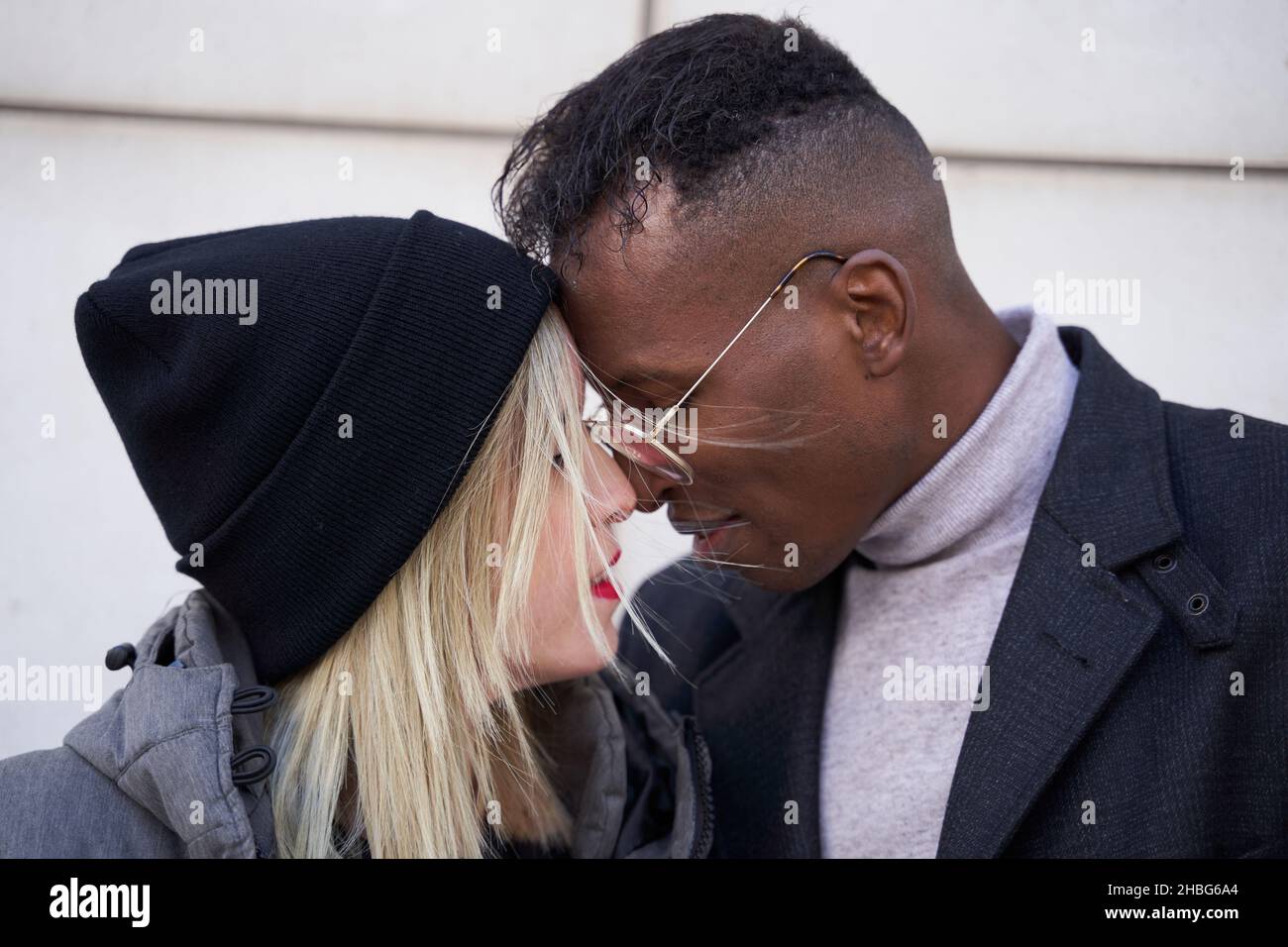 Merry African American homme et blonde femme en vêtements de dessus touchant nez et sourire tout en se tenant près du mur moderne de bâtiment, pendant la date romantique sur la rue de ville Banque D'Images