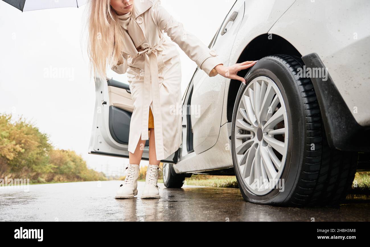 Vue rognée du bas de la femme blonde avec parapluie, garer sa voiture blanche sur le côté de la route et vérifier manuellement la résistance du pneu endommagé de la roue arrière de voiture. Banque D'Images