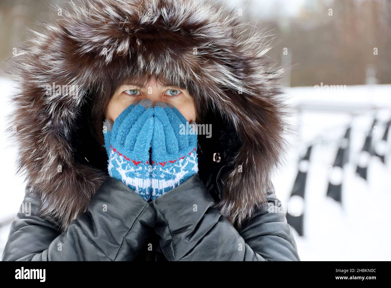 Femme en cuir manteau avec capuche en fourrure couvre son visage par les mains dans des gants tricotés debout sur une rue pendant la neige.Temps de gel en hiver Banque D'Images