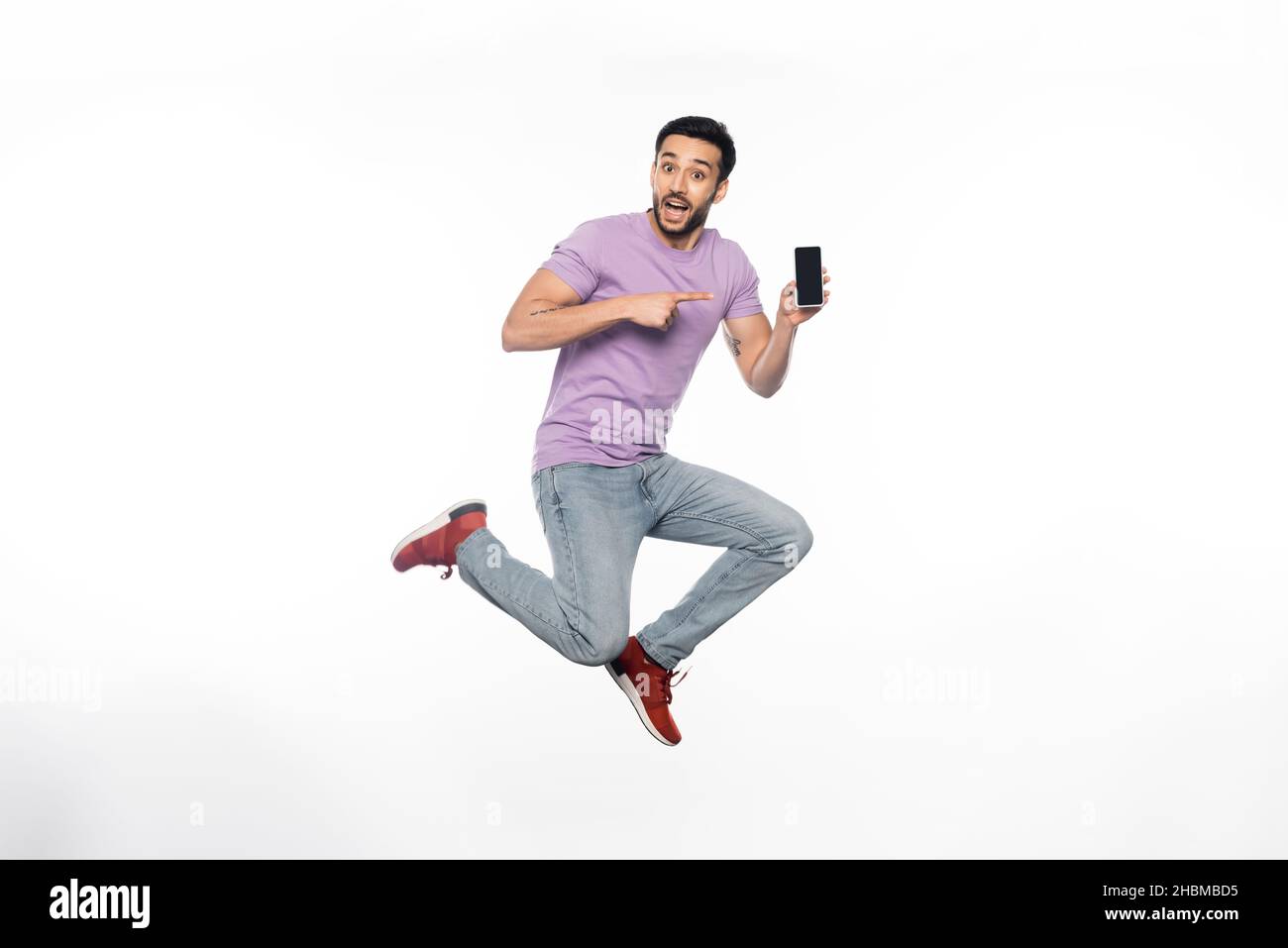 homme excité en jeans et t-shirt violet lévitant tout en pointant vers le smartphone sur blanc Banque D'Images