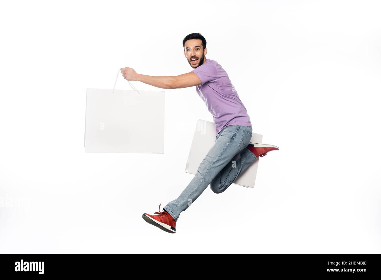 homme heureux en jeans et t-shirt violet lévitant avec des sacs de shopping isolés sur blanc Banque D'Images
