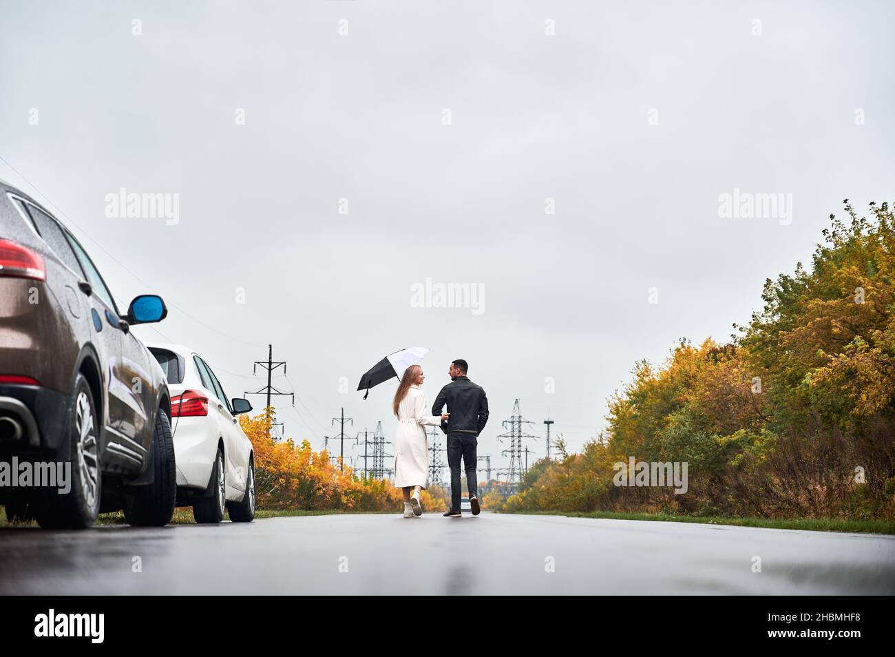 Vue arrière de la dame avec parapluie et son petit ami marchant sur la route à l'automne jour pluvieux.En toile de fond, des tours électriques en hauteur, du ciel gris et des buissons jaunes sur les routes.Deux voitures à droite. Banque D'Images