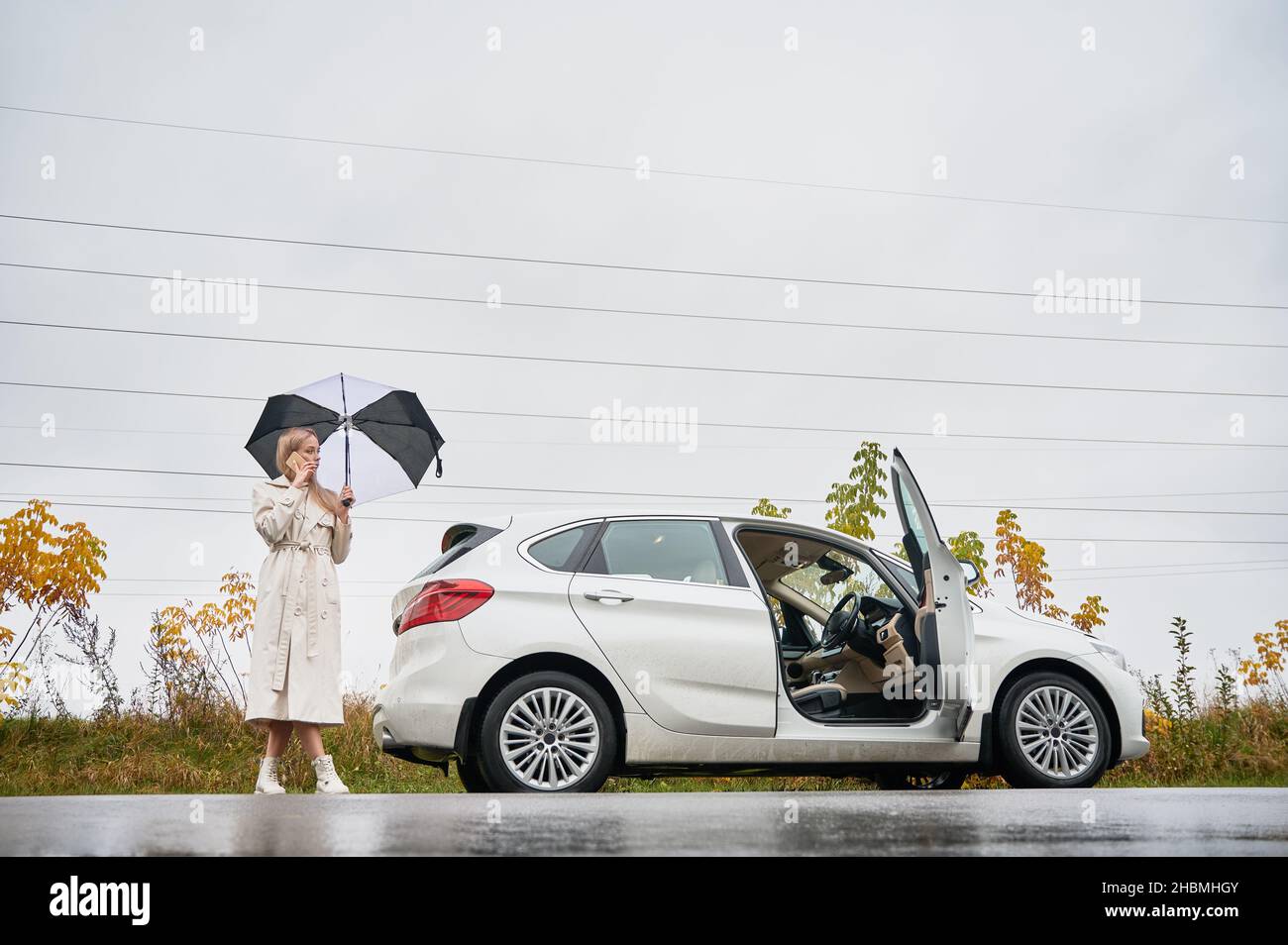 Blonde fille de parking sa voiture blanche avec porte ouverte sur le côté de la route sur la toile de fond du champ d'automne et du ciel gris.Femme avec parapluie debout derrière auto, parlant au téléphone et regardant à gauche. Banque D'Images
