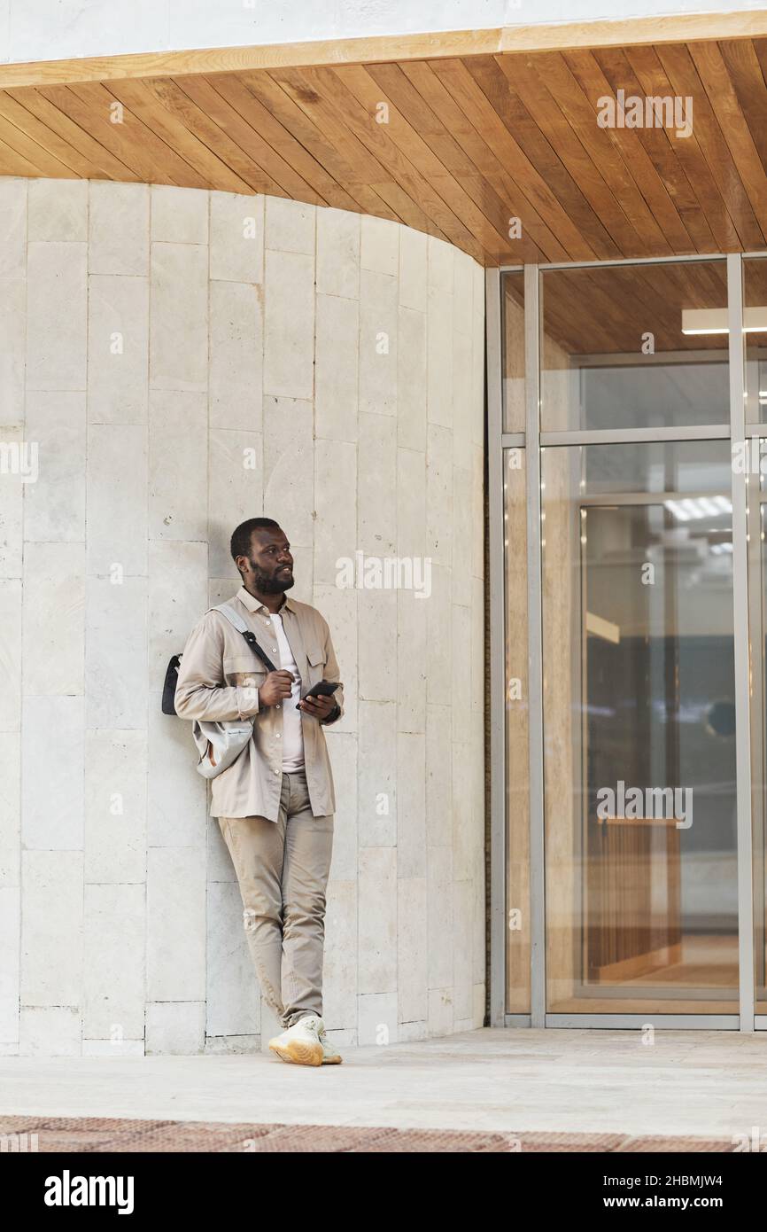 Portrait vertical complet d'un homme noir adulte utilisant un smartphone en attendant dans un immeuble de bureaux Banque D'Images