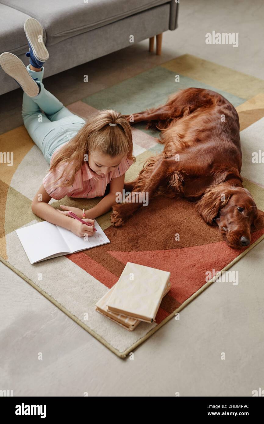 Portrait à grand angle d'une jolie fille blonde qui fait ses devoirs tout en étant couché sur le sol avec son chien Banque D'Images