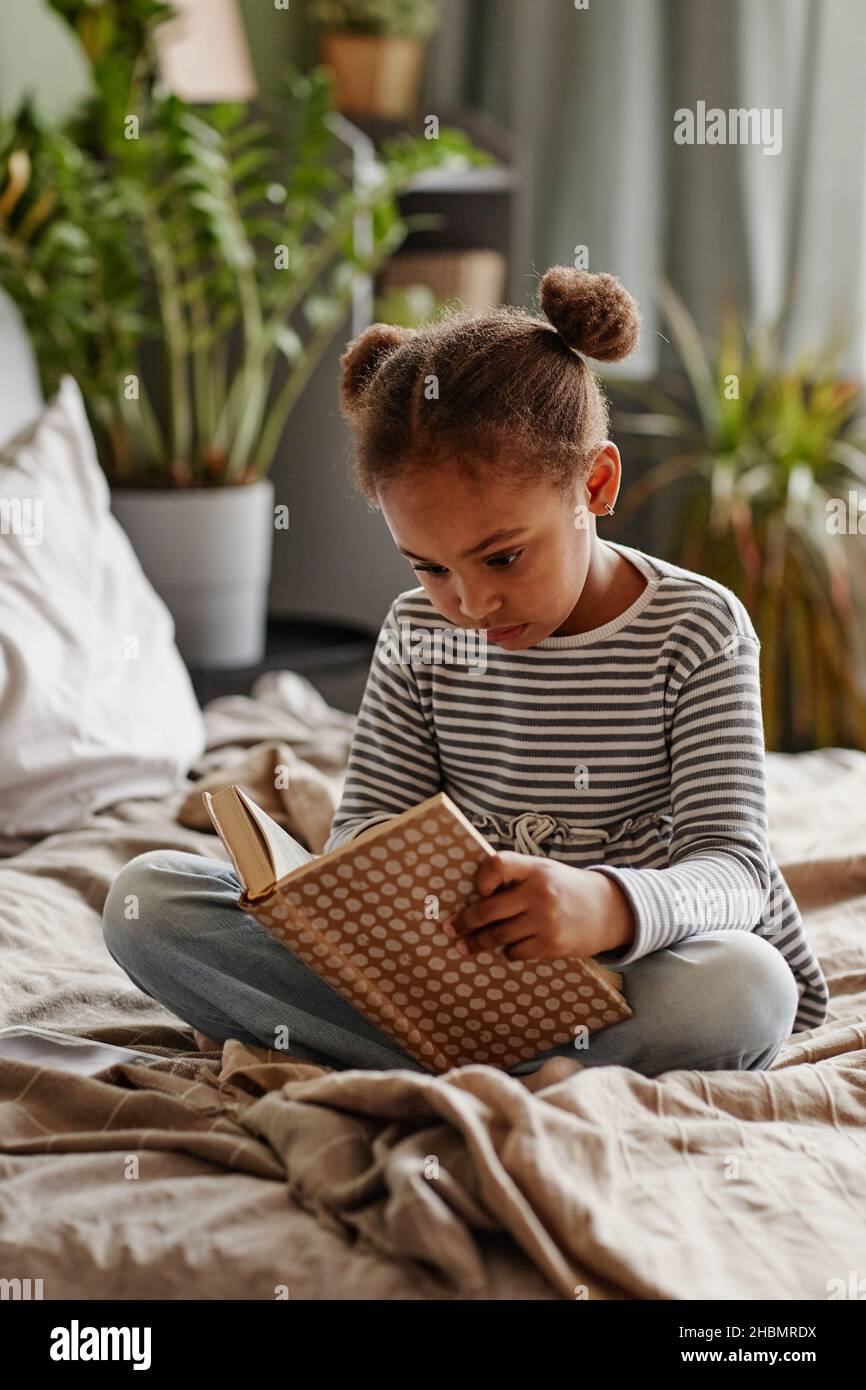 Portrait vertical du joli livre de lecture de fille afro-américaine tout en étant assis sur le lit dans un intérieur confortable Banque D'Images