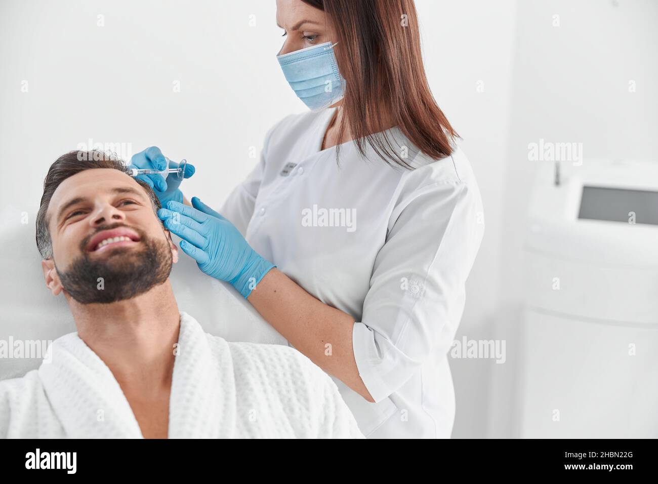 Un homme souriant avec une barbe subit une procédure de mésothérapie avec un cosmetologue professionnel en clinique Banque D'Images