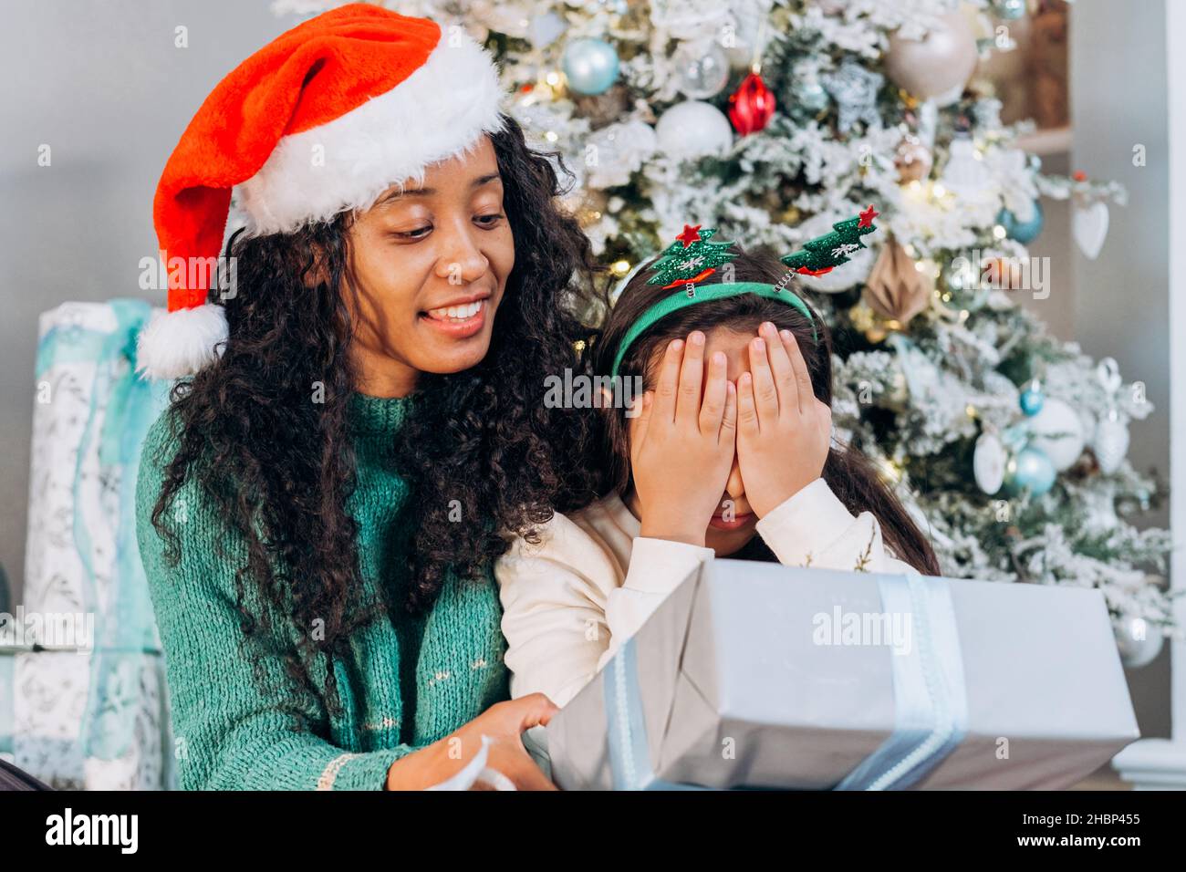 Une mère afro-américaine aux cheveux bouclés surprend sa fille brune portant un bandeau de vacances avec des cadeaux souriants près de l'arbre de Noël Banque D'Images