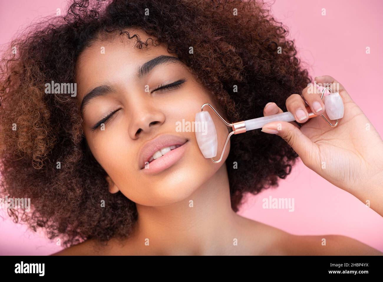 Portrait d'une jeune femme à la peau sombre et bien entretenue avec des cheveux luxuriants, faisant un massage du visage avec l'aide d'un rouleau avec les yeux fermés sur un fond rose gros plan, les soins et les soins de la peau Banque D'Images
