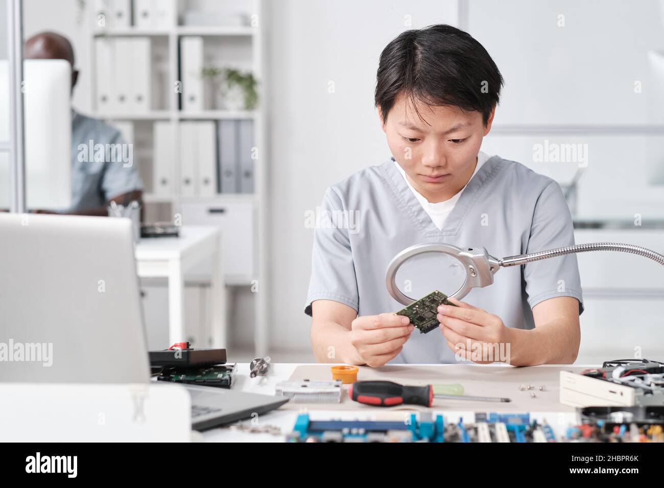 Jeune femme chinoise en uniforme regardant la puce à bord à travers la loupe tout en étant assise près du bureau Banque D'Images