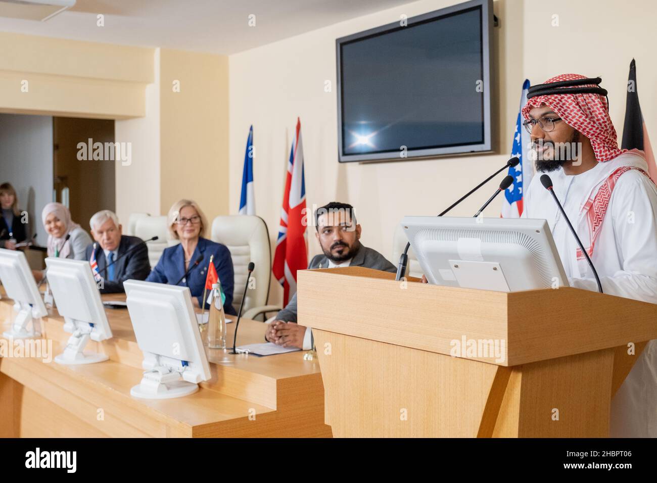 Jeune homme arabe confiant dans des vêtements traditionnels debout par tribune et parlant en micro lors d'une conférence politique Banque D'Images