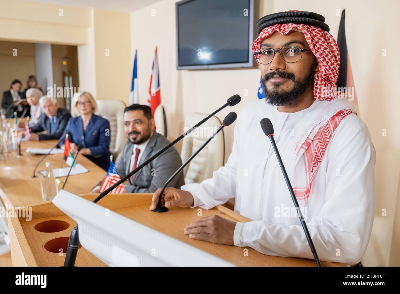 Jeune haut-parleur arabe regardant la caméra en se tenant debout près d'une tribune avec microphone sur fond de délégués étrangers Banque D'Images