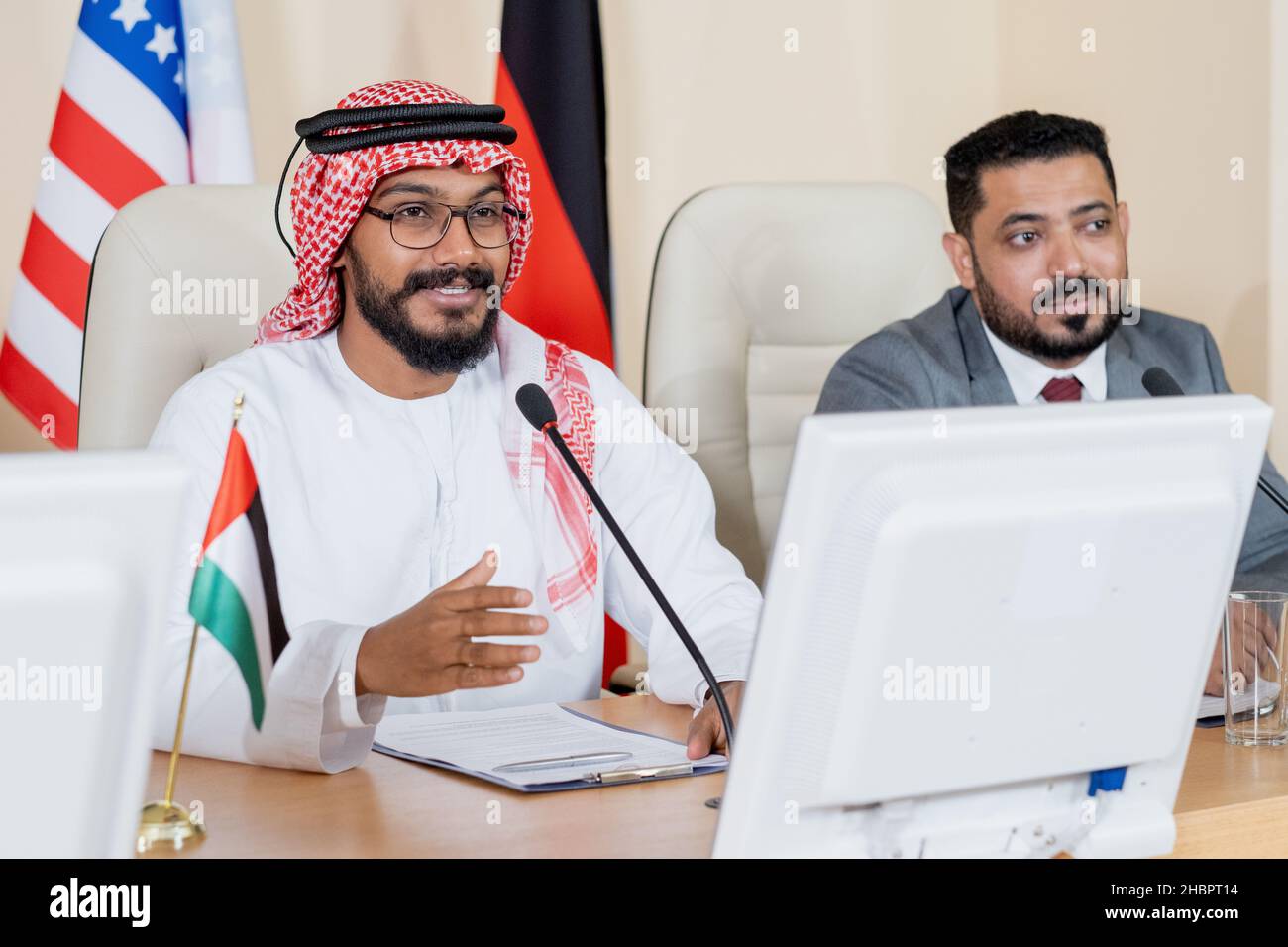 Jeune délégué arabe confiant parlant en micro lors d'une conférence ou d'un sommet politique contre un collègue étranger Banque D'Images