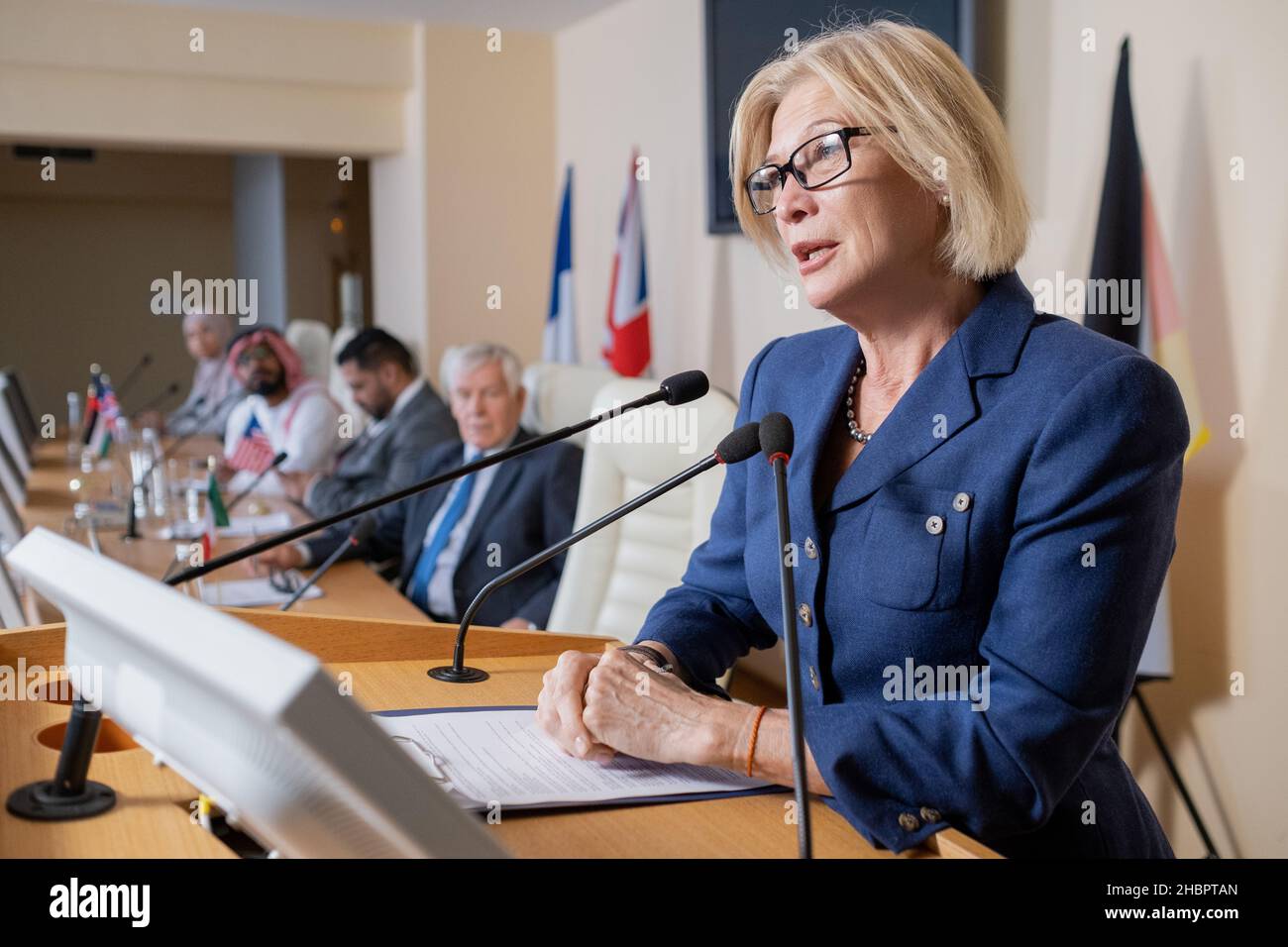 Femme blonde mature déléguée en tenue de formal parlant en micro tout en se tenant debout par Tribune au forum politique Banque D'Images