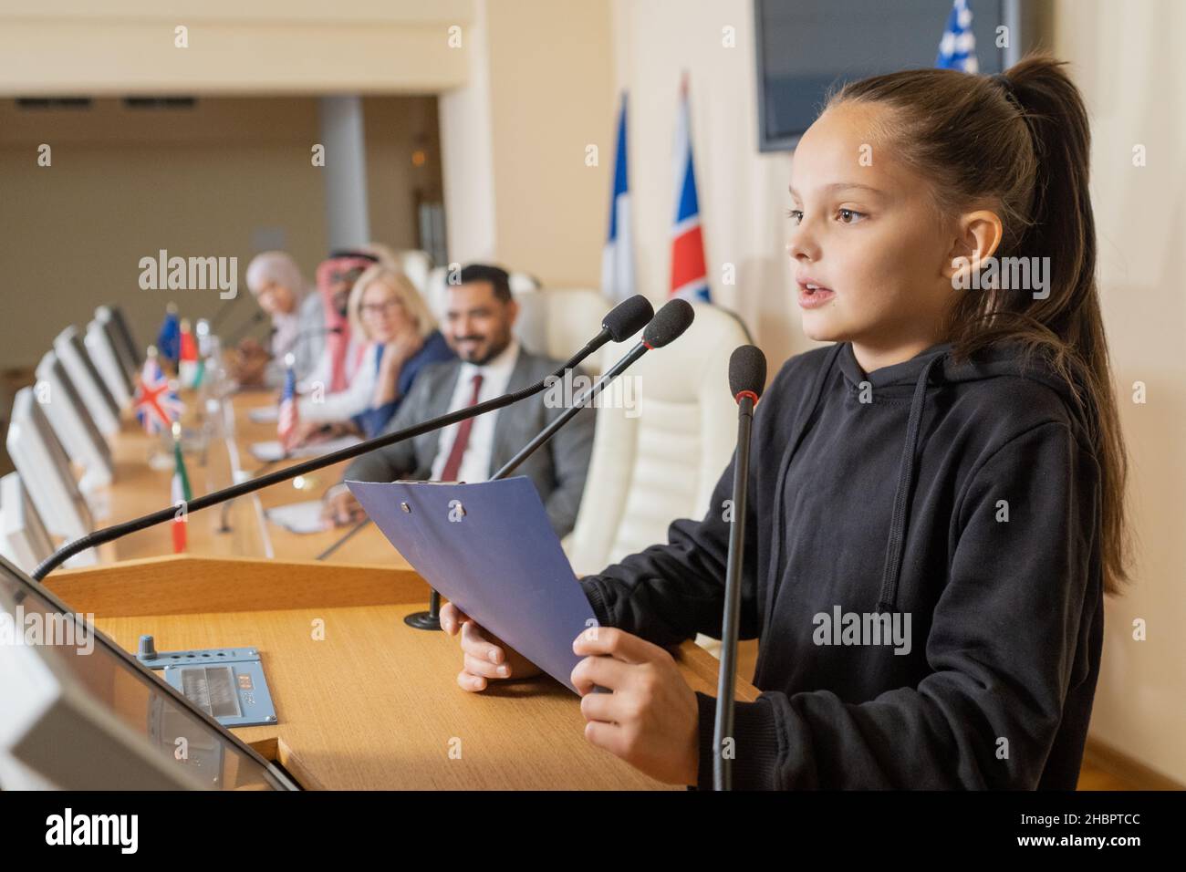 Jolie écolière avec presse-papiers parlant dans le microphone tout en faisant un rapport par Tribune contre la rangée de délégués étrangers Banque D'Images