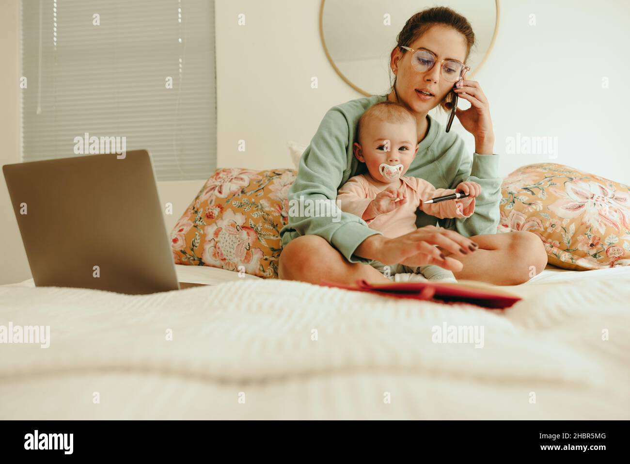 Une maman qui travaille prend un appel téléphonique tout en étant assise sur un lit avec son bébé.Une mère célibataire communique avec ses clients tout en travaillant à domicile.Nouveau maman ba Banque D'Images