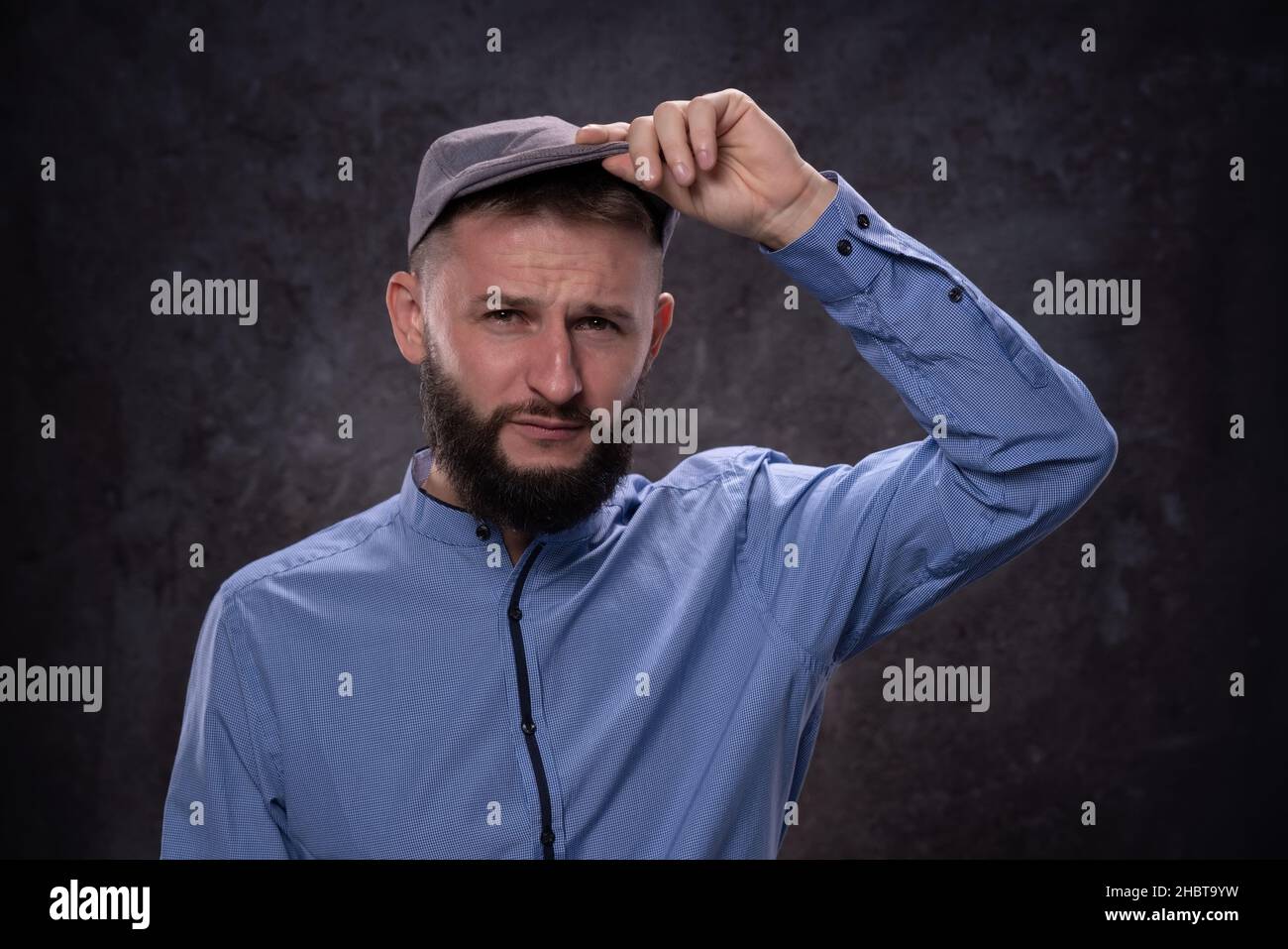 Un jeune homme européen barbu montre des émotions sérieuses étant surpris en prenant la visière d'une casquette à la mode.Portrait sur fond sombre Banque D'Images
