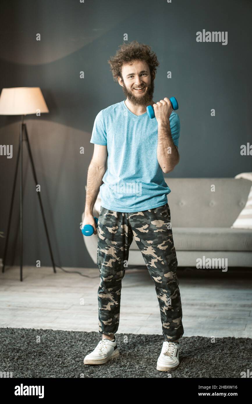 Souriant jeune homme à la maison vêtements fait ses entraînements quotidiens avec des haltères à la maison.L'homme gentil pompe ses muscles de bras.Photo de haute qualité Banque D'Images