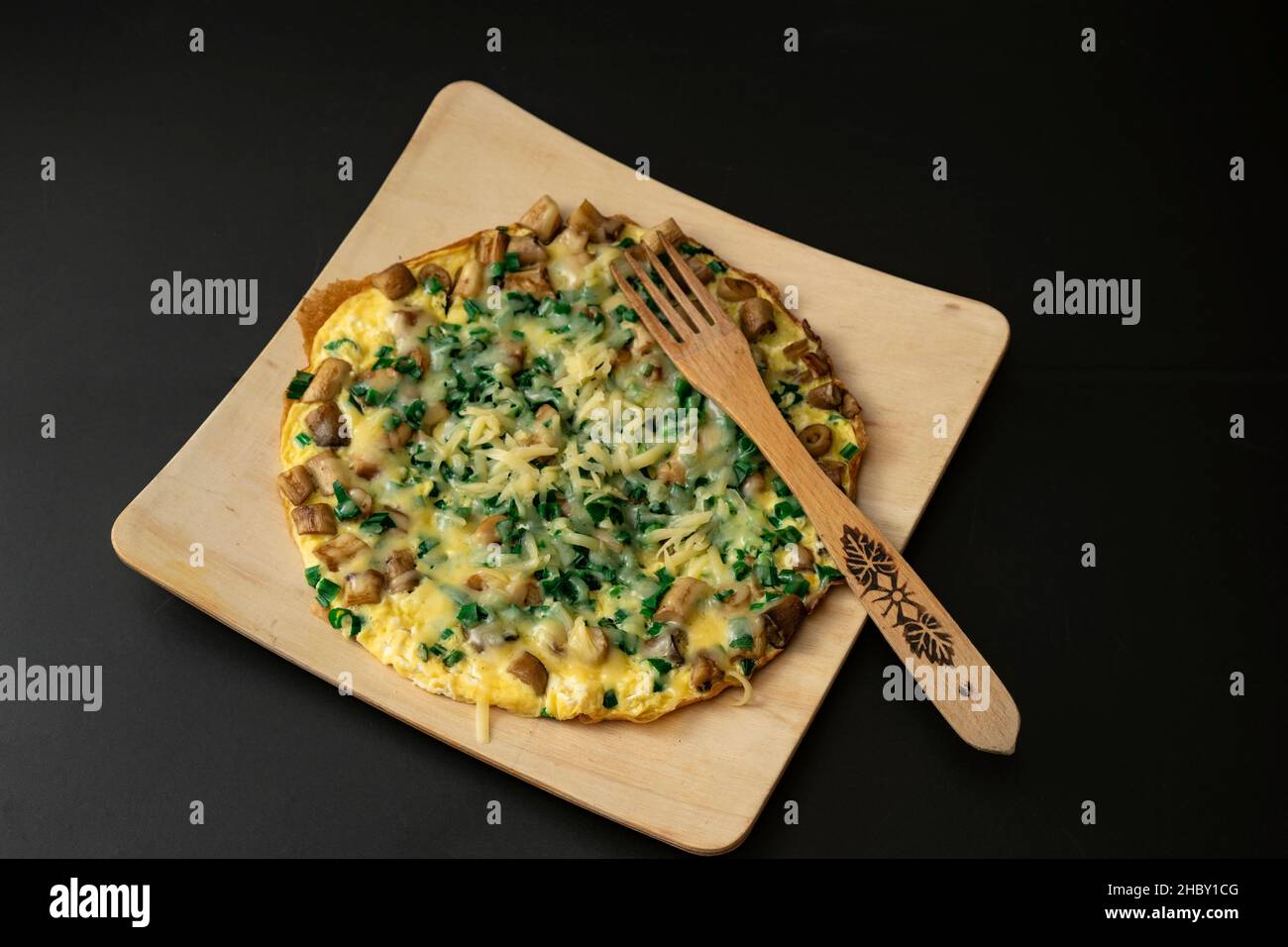 Omelette rustique aux œufs avec oignon de printemps et parmesan sur une assiette en bois avec une fourchette en bois isolée sur le noir Banque D'Images