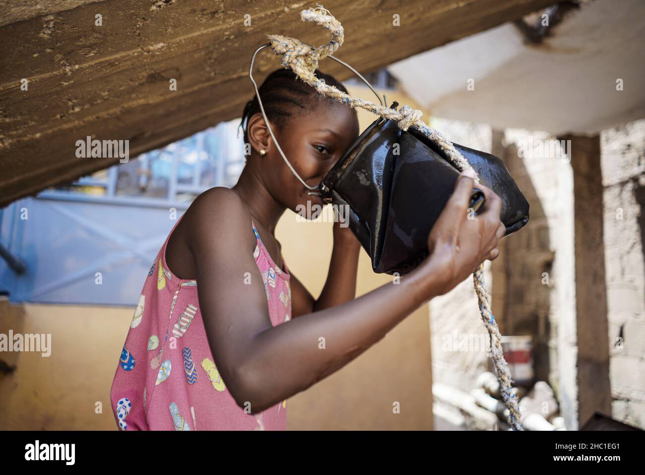 Une jeune fille africaine noire mignonne à regarder un appareil photo tout en buvant de l'eau douce à partir d'un seau en caoutchouc suspendu au toit.Concept d'approvisionnement en eau domestique Banque D'Images