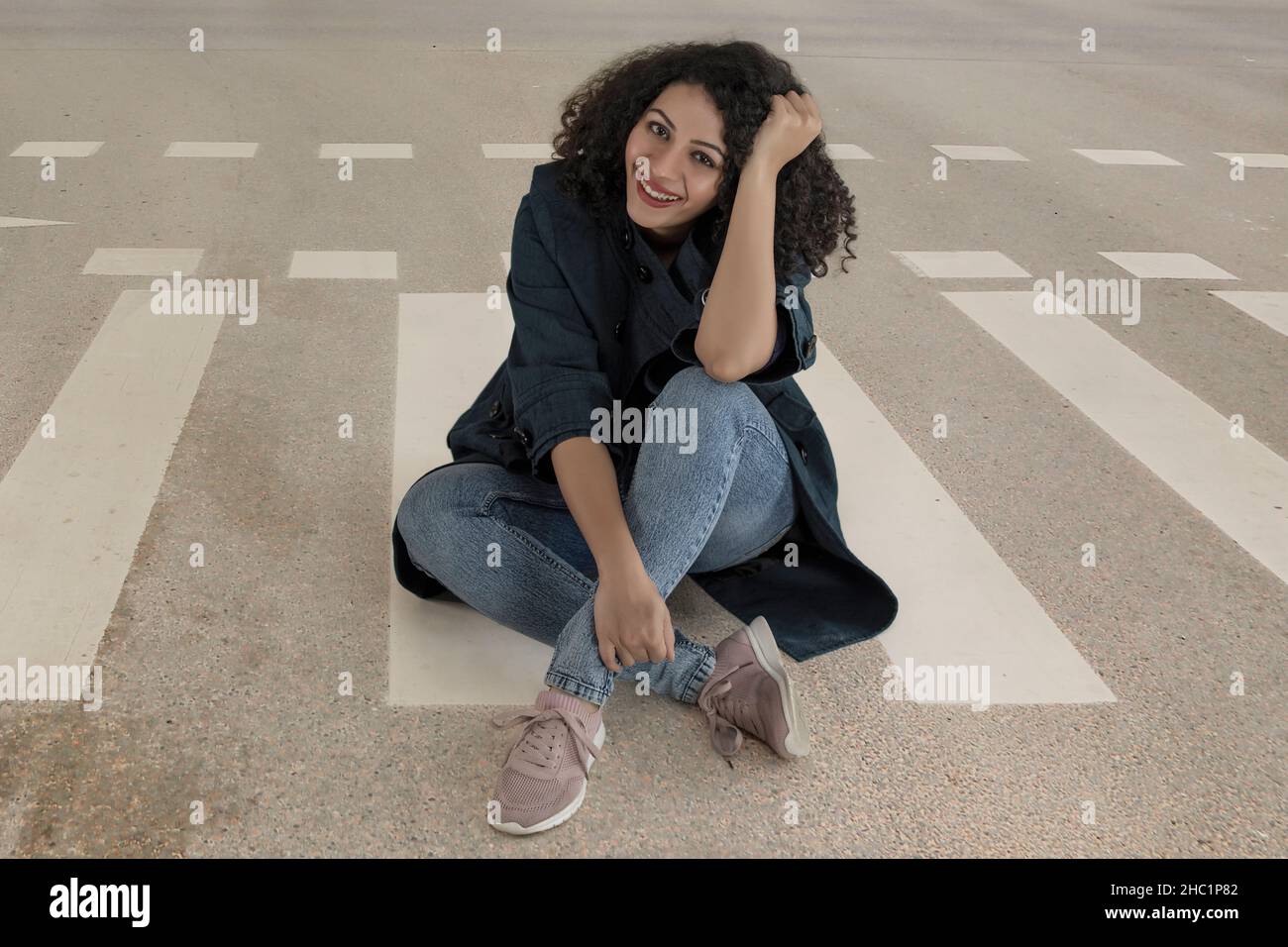Belle femme souriant et regardant l'appareil photo tout en étant assise dans la rue avec la main sur la tête Banque D'Images