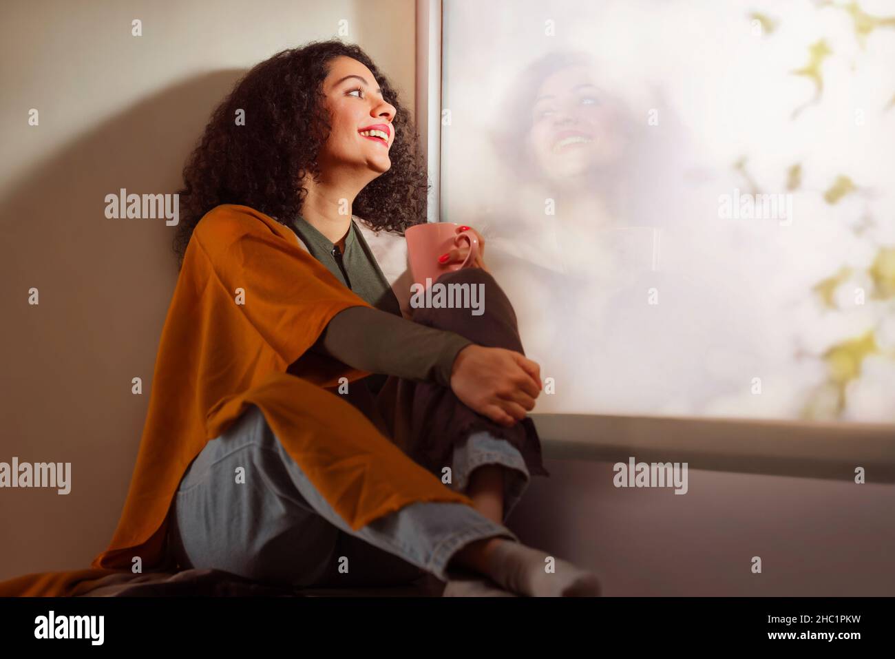 Bonne jeune femme adulte tenant une tasse de café près de la fenêtre avec réflexion dans le salon moderne Banque D'Images