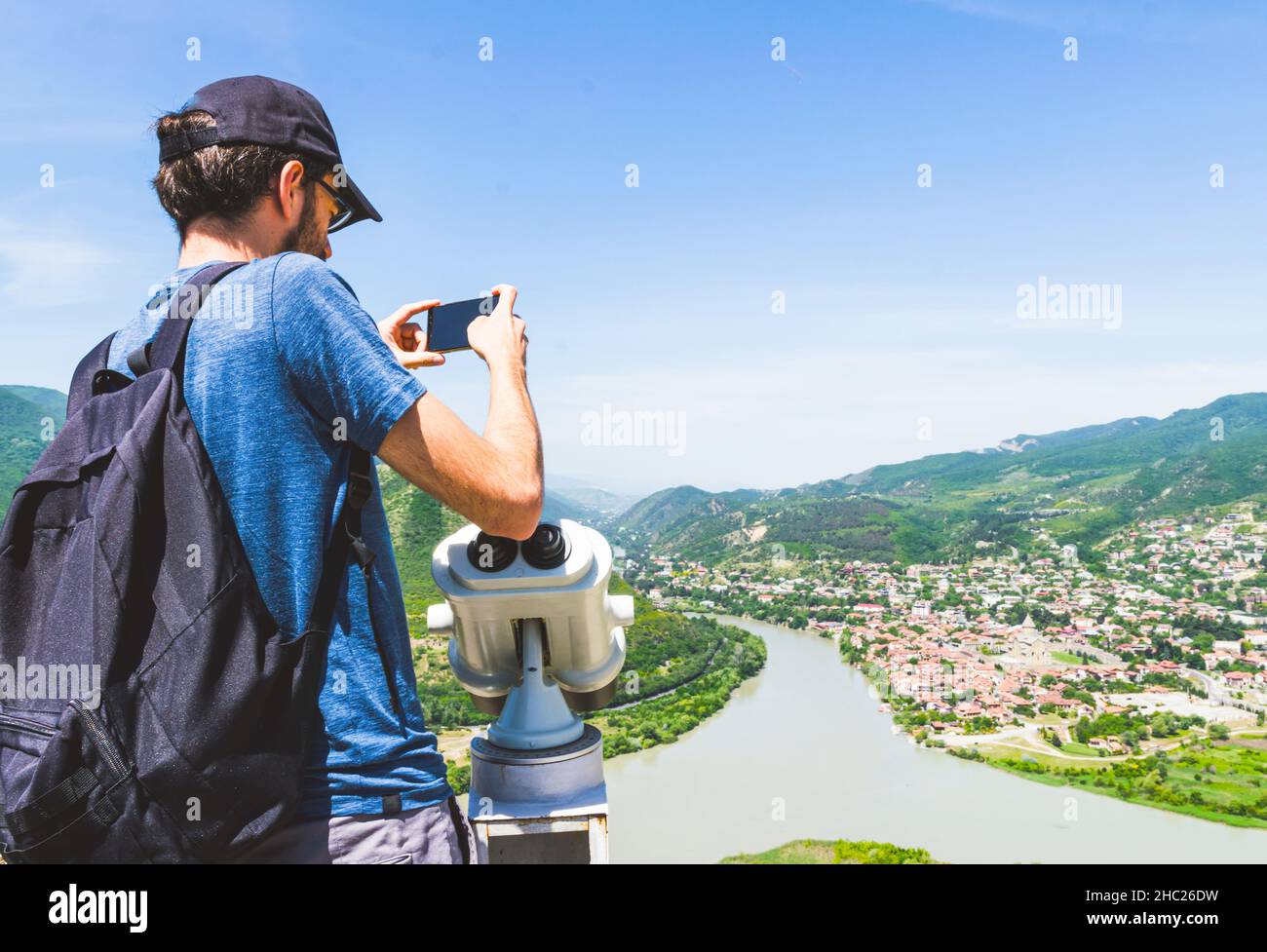Homme touriste en chemise bleue se tient près des jumelles point de vue, tient le téléphone portable et prend une photo de la ville de Mtskheta et de la rivière Mgvari.Déplacement Banque D'Images