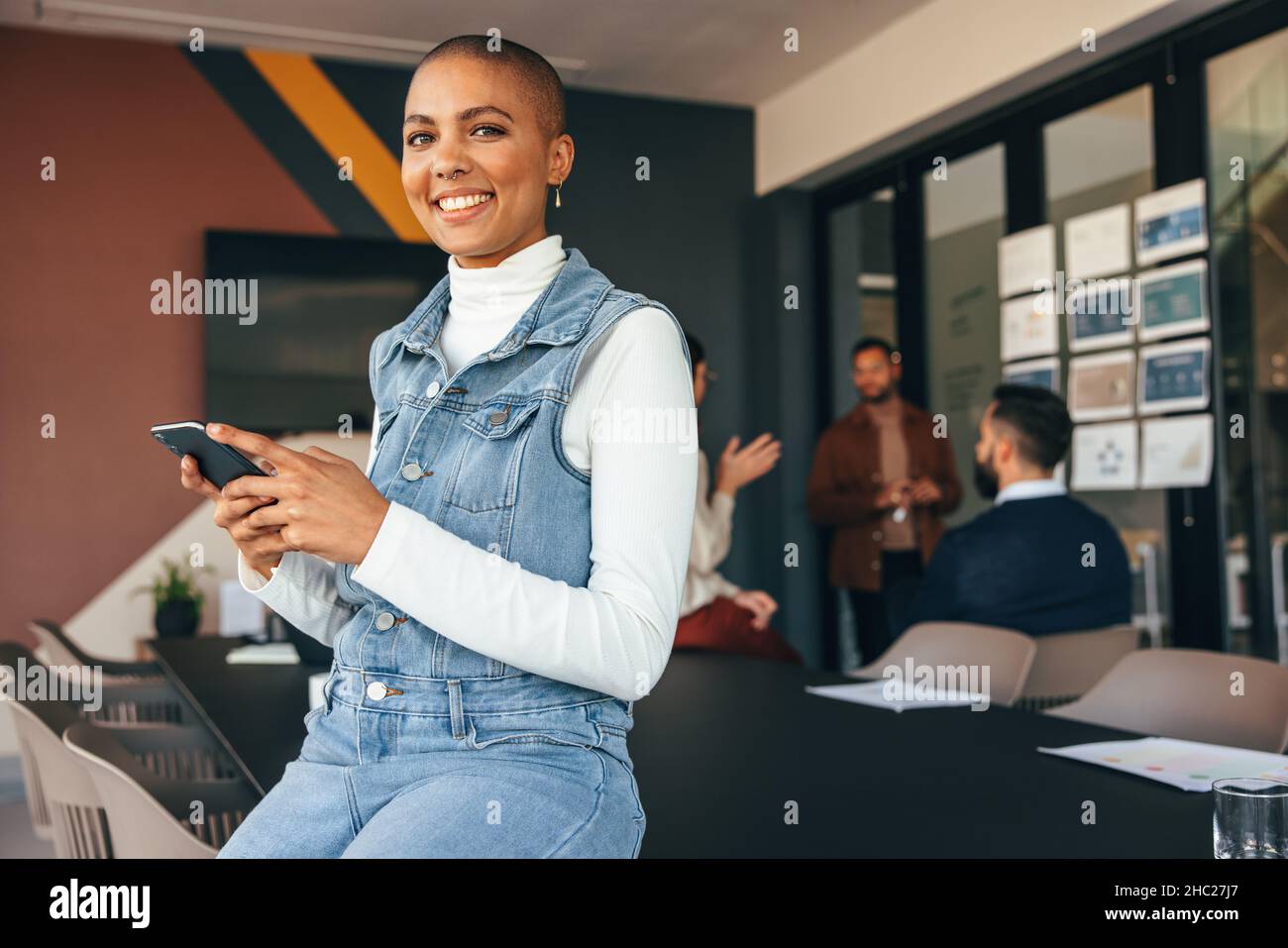 Femme d'affaires du millénaire souriant à l'appareil photo tout en tenant un smartphone.Jeune femme d'affaires gaie assise sur une table de réunion avec son collègue Banque D'Images