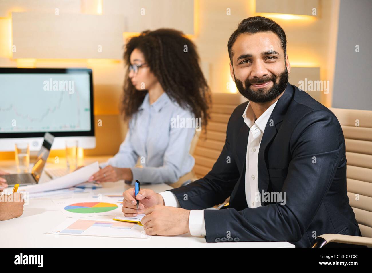 Vue portrait de l'homme d'affaires multiracial à barbe satisfait souriant à l'appareil photo tandis que la femme sortante tient des papiers et travaille dans son lieu de travail à l'intérieur Banque D'Images