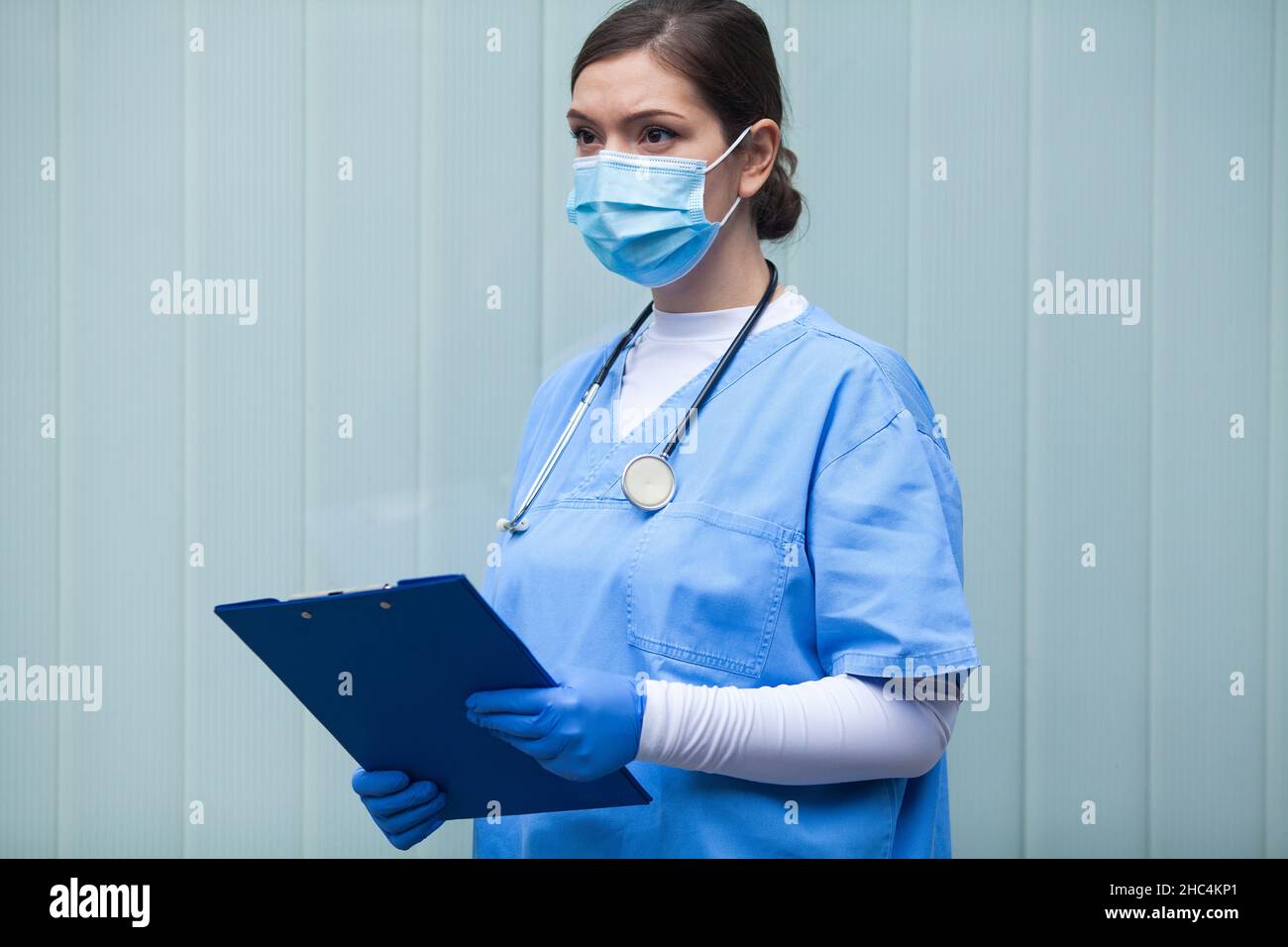 Portrait de jeune femme de race blanche médecin NHS, debout devant le mur de verre bleu, regardant loin, portant l'équipement de protection bleu, fa Banque D'Images